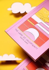 Description: Copies of “The Land of Uncertainty” sit on a yellow table with paper clouds. The cover is pink, yellow, and red with various abstract shapes, showing a raised texture to the cover.