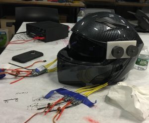 Description: An adapted motorcycle helmet with colorful electrical wires coming out of it sits on a workbench.